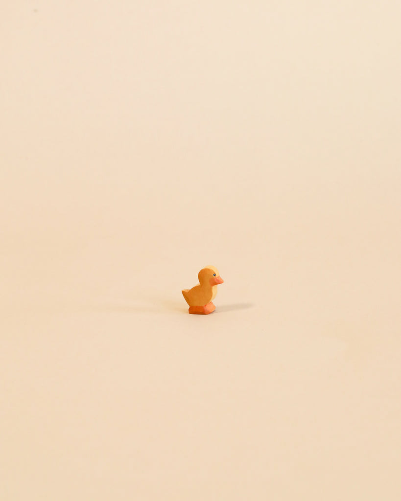 A small, orange Ostheimer Duckling centered on a plain, light beige background. The duck casts a soft shadow behind it, emphasizing its isolated position and evoking the charm of handcrafted wooden toys.