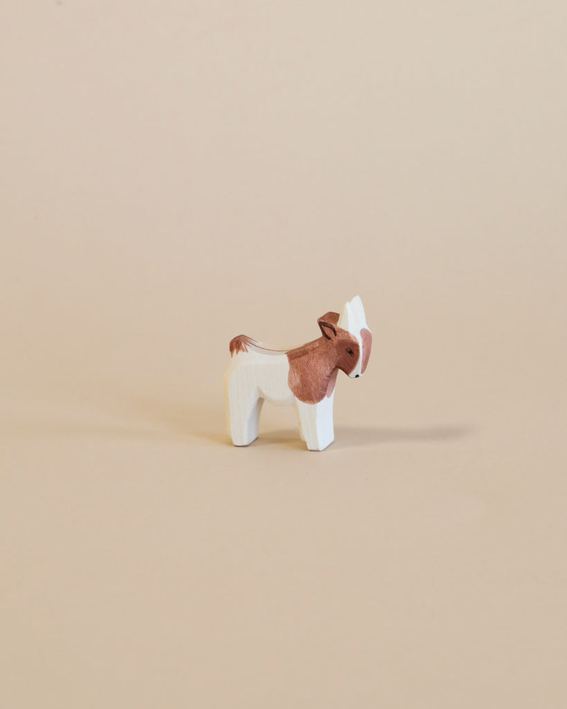 A small handcrafted Ostheimer Goat Kid - Standing with a brown body and white face and legs, standing against a plain beige background.