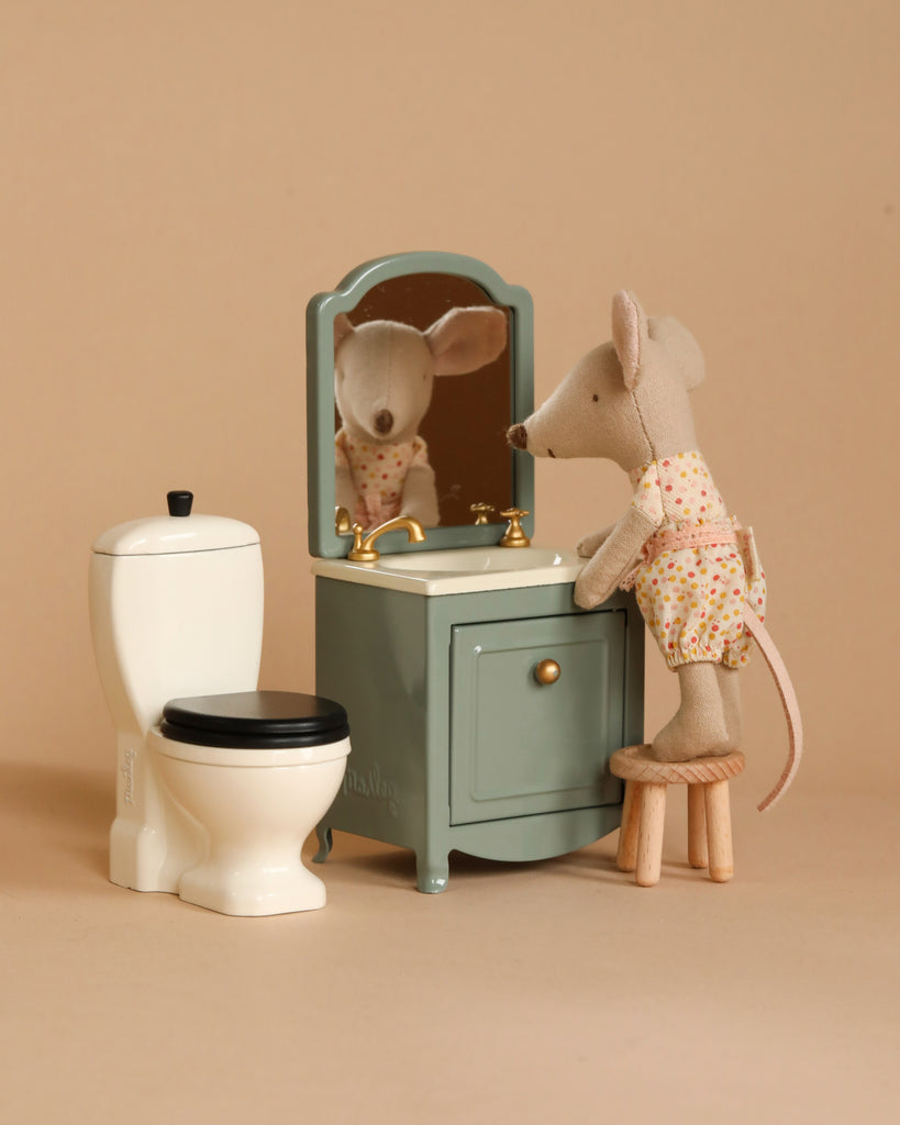 A Maileg little sister plush mouse toy, dressed in a colorful patterned outfit, stands on a stool in front of the Maileg Bathroom Starter Set which includes a miniature vanity with a mirror and sink, along with a white toilet beside the vanity. The background is a plain beige color. This charming children's toy set brings imaginative play to life.