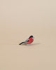 A small, colorful handcrafted Ostheimer Bullfinch with a pinkish-red chest and grey-black wings, standing isolated against a plain, light beige background.