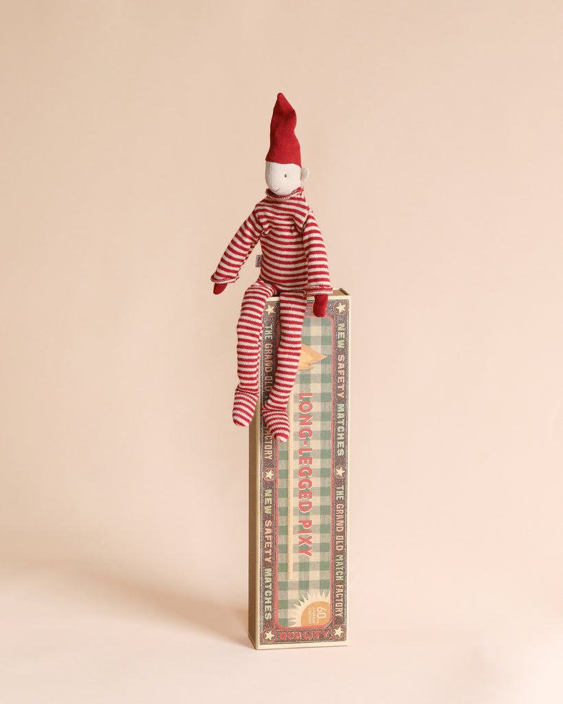 A Maileg Long Legged Pixy wearing red and white striped pajamas and a red hat is sitting on top of an upright, vintage-style rectangular box with decorative matchbox text and patterns. The background is a soft, light beige color.