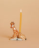 The Giraffe Cake Topper, crafted from fine porcelain and adorned with natural giraffe markings, serves as a charming candle holder. It features a single lit yellow-orange candle positioned on its back. This heirloom-quality piece is set against a plain beige background, making it a collectible keepsake for any home.