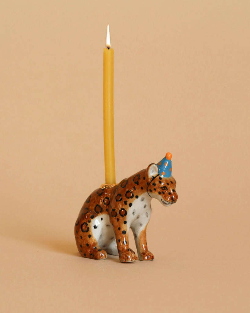 A hand-painted ceramic Jaguar Cake Topper features a jaguar adorned with a blue party hat with a white pompom, functioning as a candle holder. A tall, lit yellow candle is elegantly placed on its back, making this collectible keepsake stand out against the plain beige background.