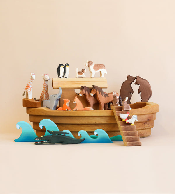 A collection of Ostheimer Handmade Large Wooden Ark, including giraffes, bears, and penguins, artistically arranged on and around a wooden boat against a peach background.