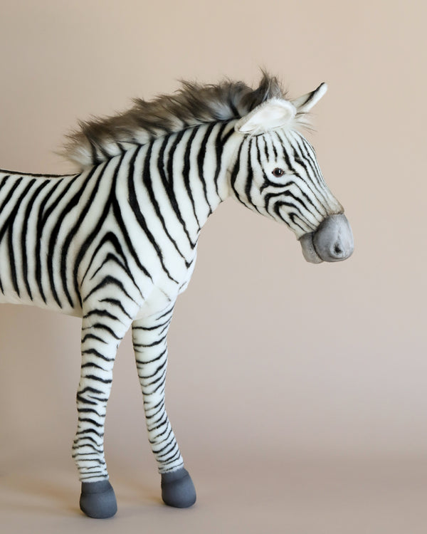 The Large Zebra Stuffed Animal, featuring black and white stripes, stands against a neutral beige background. This hand-sewn zebra showcases a fluffy mane and gray hooves, facing to the right—embodying the essence of realistic plush animals with its distinctive character.