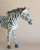 The Large Zebra Stuffed Animal, featuring black and white stripes, stands against a neutral beige background. This hand-sewn zebra showcases a fluffy mane and gray hooves, facing to the right—embodying the essence of realistic plush animals with its distinctive character.