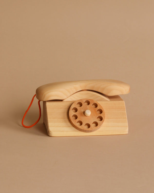 The Grimm's Wooden Telephone, a small, non-toxic toy phone with a rotary dial design, sits on a beige background. Featuring a rounded receiver and a single red string attached to the side, this charming toy is constructed from smooth, light-colored wood and boasts a simple and classic appearance.
