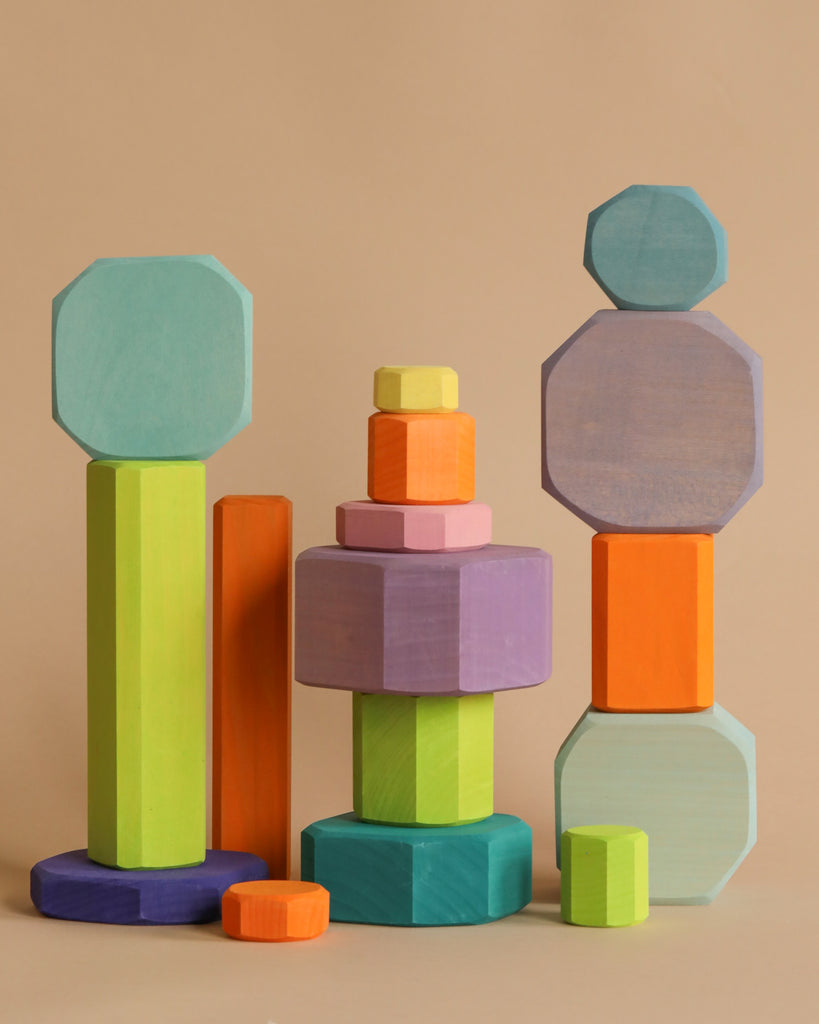 A neatly arranged stack of Grimm's Tree Slices Building Blocks, featuring vibrant, non-toxic wooden pieces in various geometric shapes like cylinders, prisms, and octagons, stands out against a plain beige background. The blocks showcase bright colors such as green, orange, purple, pink, blue, and yellow—perfect for young children.