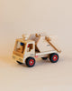 A Fagus Wooden Skip Truck with a natural finish sits on a light beige surface. The handcrafted truck has a square cab with a red, blue, and white figurine visible inside. It features red wheels with black tires and a boxy back section, suggesting it might be made in Germany.