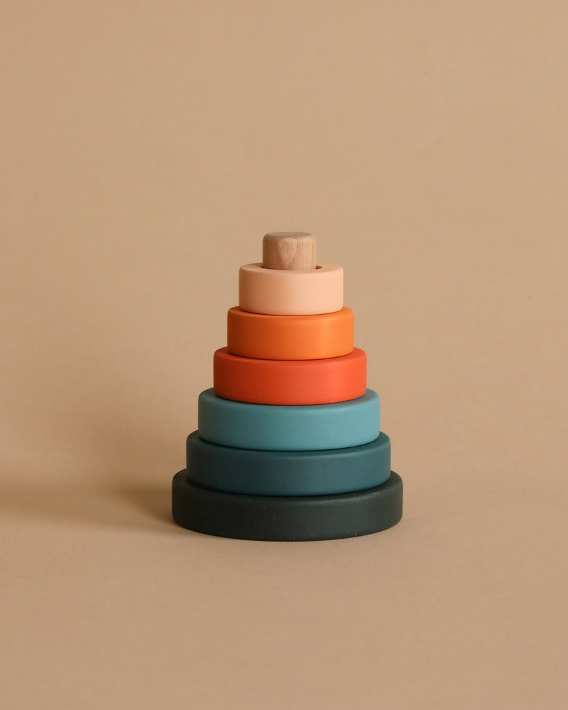 The Mini Wooden Pyramid Stacker - Tropics by SABO Concept is a handmade toy featuring circular rings in various earth-toned colors, arranged in ascending order from largest to smallest against a beige background. Painted with non-toxic paint, the rings showcase shades of dark green, blue, red, orange, and light peach.