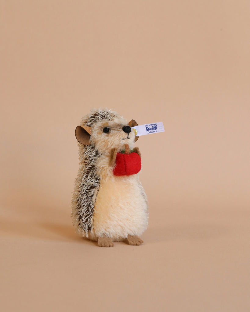 A plush Steiff, Ivo Hedgehog with Felt Apple, 4 inches toy with a fluffy belly and spiky back, holding a red apple and a small white sign with the word "sticker" in blue text, against a soft.