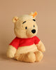 The Steiff Disney's Winnie the Pooh Bear Stuffed Plush Toy, standing 11 inches tall, features light brown fur and wears a red shirt. The plush toy sits upright against a plain beige background, sporting a small smile and black eyes. A yellow tag can be seen attached to its ear.