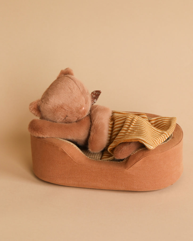 A small, brown stuffed animal resembling a bear is nestled in the Maileg Cozy Bed, with a striped yellow and white soft blanket partially covering it. The background is a neutral beige color.