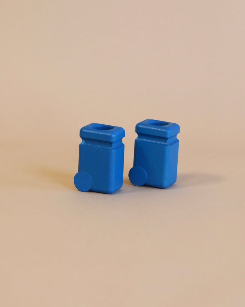 Two small Fagus Wooden Recycling Cans in blue, each resembling a miniature garbage truck or wheeled container, are set against a plain beige background. Both have a square top opening and a round, protruding wheel-like feature on one side near the base.