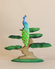 A *Handmade Wooden Peacock On A tree* with a blue head and vibrant green tail feathers stands on the branch of a stylized, multi-tiered lime wood tree with rounded green leaves. The handmade bonsai-like tree has a natural wood trunk and is painted using non-toxic paints against a plain, beige background.