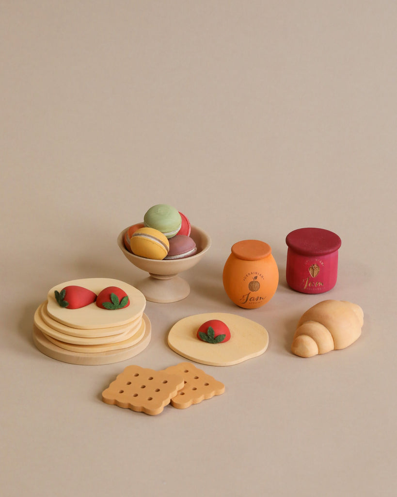 A spread of Sabo Concept Handmade Wooden Bakery Set featuring a stack of pancakes, a croissant, biscuits, jars of jam, and a bowl with colorful macarons. The setup is perfect for pretend play food on a neutral beige background.