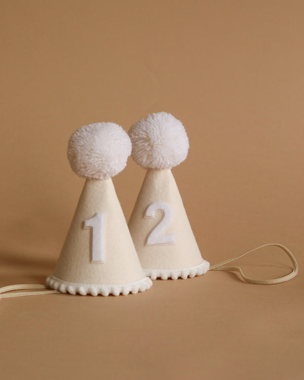 Two small White Party Hats with white pom-poms on top are displayed on a beige background, perfect for birthday celebrations. The cream-colored felt hats, adorned with zigzag trim at the base, feature numbers "1" and "2." Ideal for dress-up fun!