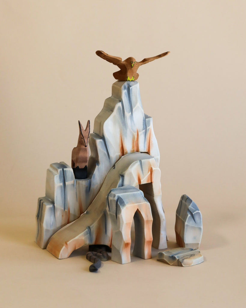 A Handmade Wooden Mountain Cliff Scene features a gray and orange colored, handmade mountain scene with a slope, a perched bird of prey at the top, and a smaller bird figure on a lower rocky ledge. Painted with non-toxic paints, small wooden rocks are scattered at the base, perfect for a Waldorf inspired play space.