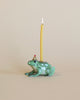 A hand-painted ceramic Frog Cake Topper with a lit yellow taper candle on its back, set against a neutral beige background. The frog has a playful expression and detailed green glazing.