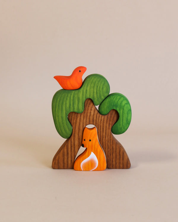 A Handmade Wooden Tree with Fox, handcrafted from ash wood and finished with water-based paint, features a whimsical tree with green foliage and a brown trunk. A bright orange bird perches on top, while an orange fox with white markings rests against the base. The backdrop is plain light beige.