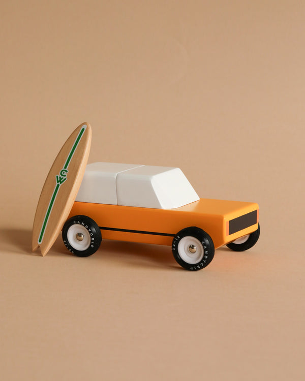 The Candylab Cotswold, a wooden toy vehicle with a minimalist design, is painted in orange and white and positioned against a beige background. Leaning against the car is a small, wooden surfboard featuring a green stripe along its length, adding a playful, beach-themed element to the scene.