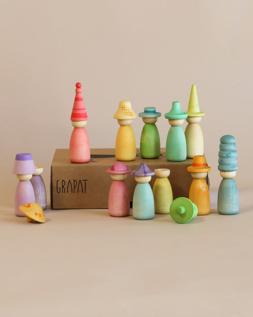 A collection of colorful, sustainable wooden toys arranged on a surface, featuring various shapes and sizes, some resembling people with interchangeable hats. The box they are on reads "Grapat Fancy Nins.