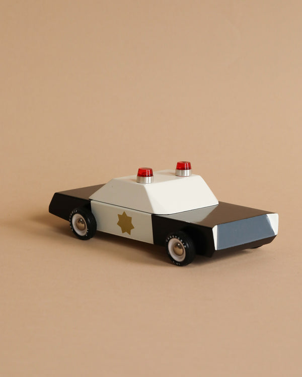 The Candylab Police Cruiser, a black and white toy police car crafted from solid Beech wood, is positioned against a beige background. It features red lights on top, rubber tires, and a gold star emblem on the side.