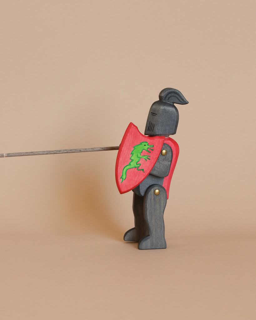 The Ostheimer Black Knight, a small wooden figurine, stands holding a lance and a red shield adorned with a green dragon emblem. Painted in dark gray with gold accents and draped in a red cloak, this knight is perfect for imaginative play and stands against a plain tan background.