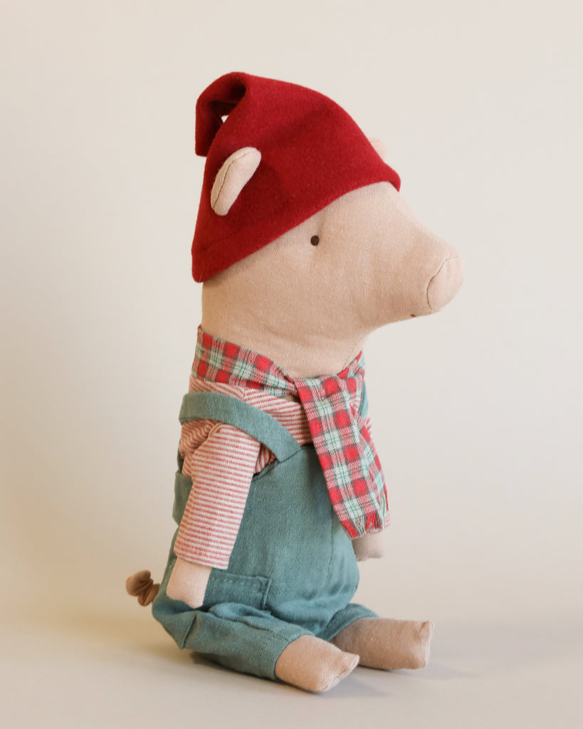 The Maileg Christmas Pig - Boy sits upright against a plain background. Dressed in a festive red hat, a green and red plaid scarf, a striped shirt, and green overalls, the pig faces to the right with its tiny tail visible at the back. This charming figure makes an adorable holiday decoration.