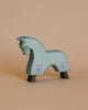 On a beige surface stands the Ostheimer Blue Horse for Knight, a light blue, handcrafted wooden horse figurine with simple features. It boasts two small shields adorned with a fleur-de-lis emblem painted on its sides and has dark-colored hooves. The solid beige background sets the stage for imaginative play inspired by classic Ostheimer toys.