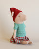 The Maileg Christmas Pig - Girl, perfect for the holiday season, is dressed in a red hat, a green knitted sweater, and a red plaid skirt while sitting against a neutral background.