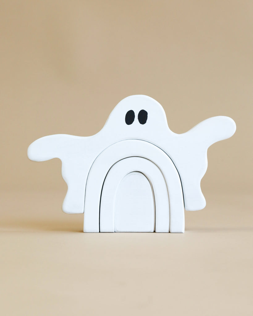 A minimalist wooden toy in the shape of a ghost is painted with Non-Toxic wood dye and features black eyes. Handcrafted from poplar hardwood, the Handmade Wooden Ghost Stacker consists of three arch pieces that stack together to form the ghost's body and arms. The background is a plain beige color.