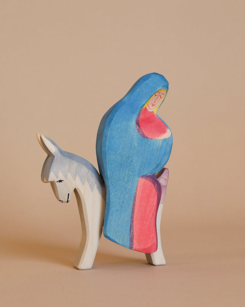 The Ostheimer Mary on Donkey is a small wooden figurine depicting a person in a blue and red cloak riding a white donkey. The art style is minimalist, with bold colors and smooth lines. Perfect for imaginative play, this piece reflects the timeless charm of handcrafted toys against a plain, light beige background.