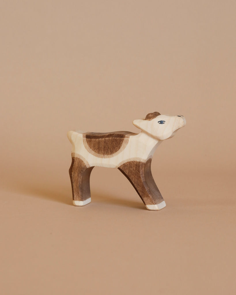 An Ostheimer Reindeer Calf figurine, handcrafted from wood, stands against a plain beige background. This piece inspired by Ostheimer toys features painted details in light brown, dark brown, and white hues with small blue eyes—perfect for imaginative play.