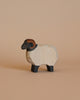 A small handcrafted Ostheimer Moorland Ram standing on a beige background. This wooden figurine features a cream-colored body, black legs, and a black face with a distinctive brown marking around its head. Perfect for imaginative play, the carving details give its wool a textured appearance.