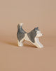 The Ostheimer Sled Dog - Running is a small, wooden figurine with a playful stance, painted in white and gray hues. This handcrafted piece features its front legs extended forward and its tail raised, capturing a stretching or playful pose. The background is a plain, light brown color.