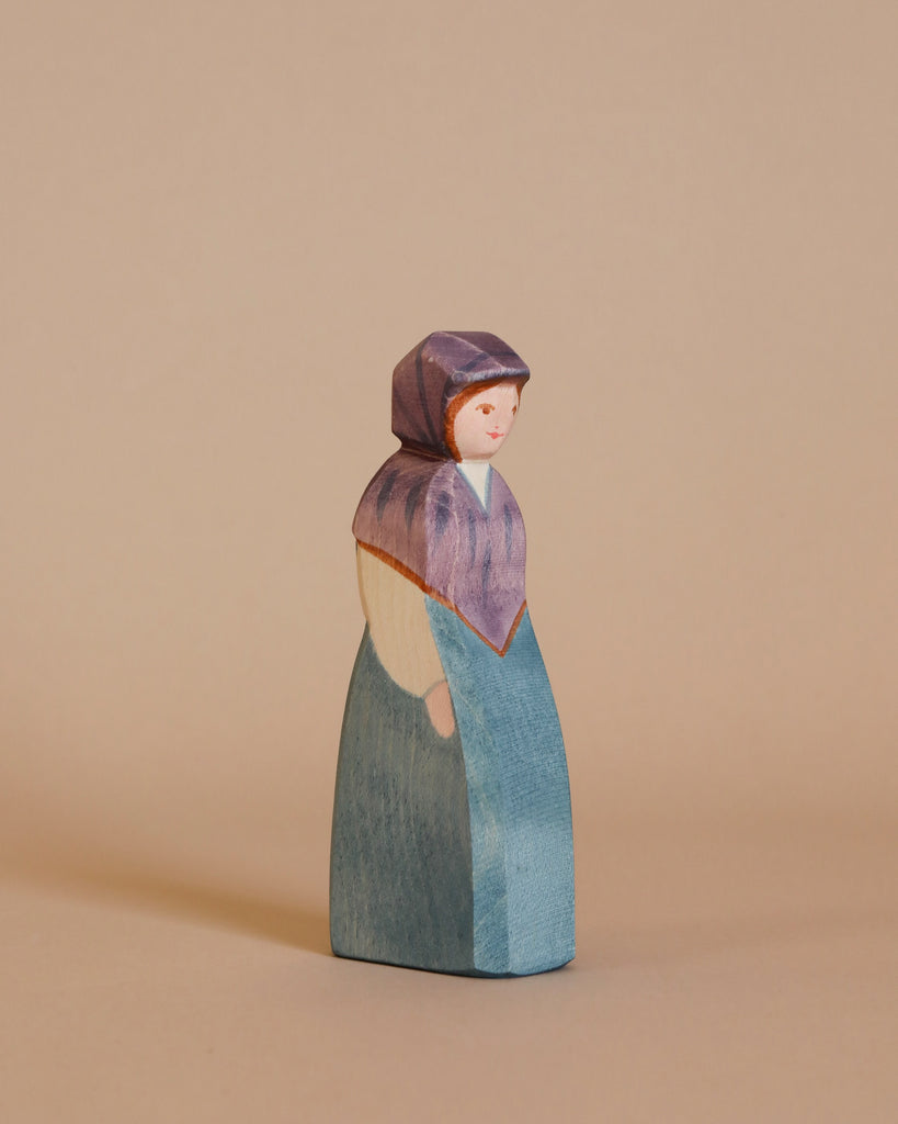 The Ostheimer Market Woman is a handcrafted wooden figurine featuring a woman adorned in a purple headscarf and blue dress, set against a plain beige background. The figurine boasts minimal facial features and a solid, blocky shape, perfect for imaginative play while capturing the timeless charm of Ostheimer wooden toys.