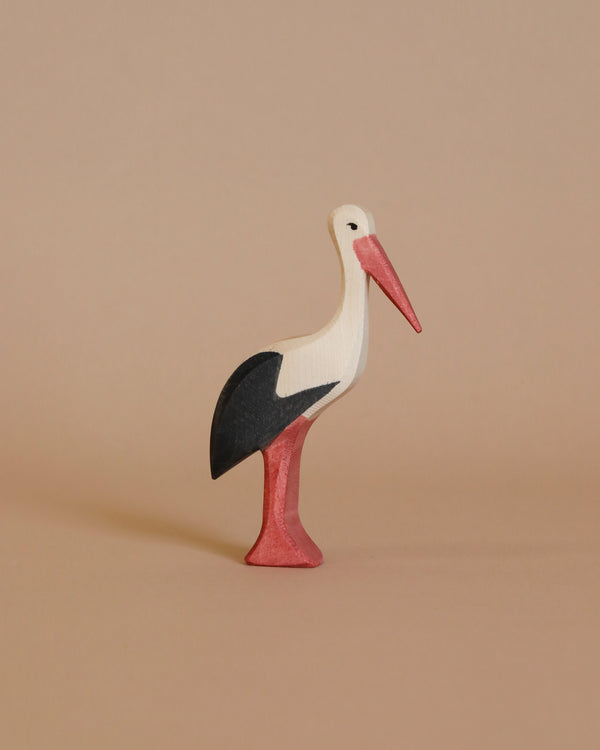 An Ostheimer Stork wooden figurine stands on a light brown surface, showcasing its handcrafted elegance. The stork, with its white head and body, black wings, and red beak and legs, invites imaginative play against the neutral background. This piece perfectly embodies the charm of Ostheimer toys.