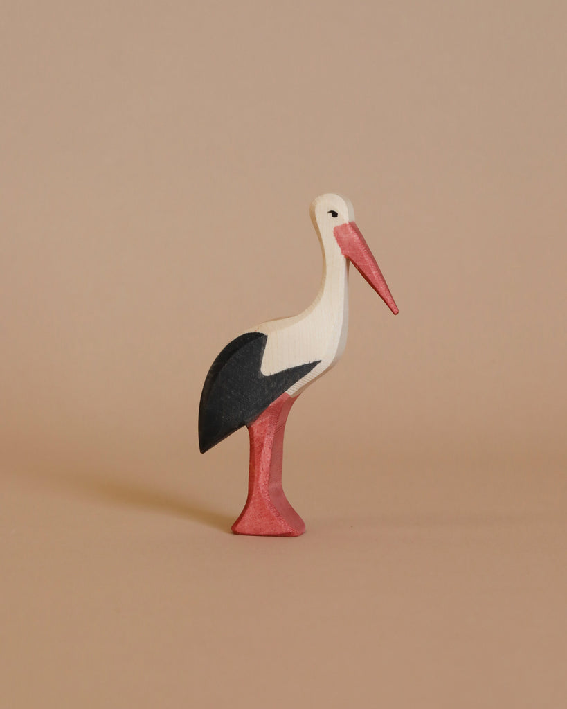 An Ostheimer Stork wooden figurine stands on a light brown surface, showcasing its handcrafted elegance. The stork, with its white head and body, black wings, and red beak and legs, invites imaginative play against the neutral background. This piece perfectly embodies the charm of Ostheimer toys.