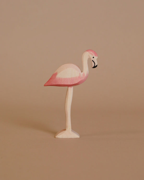 The Ostheimer Flamingo, a small wooden figurine handcrafted from sustainable materials and painted in shades of pink, stands gracefully on one leg against a plain beige background. Its body is primarily pink with subtle white accents and features a black-tipped beak. The overall design is simple and minimalist, echoing the classic charm reminiscent of Ostheimer toys.