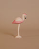 The Ostheimer Flamingo, a small wooden figurine handcrafted from sustainable materials and painted in shades of pink, stands gracefully on one leg against a plain beige background. Its body is primarily pink with subtle white accents and features a black-tipped beak. The overall design is simple and minimalist, echoing the classic charm reminiscent of Ostheimer toys.