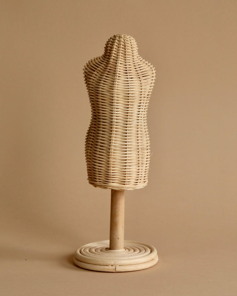 In a cozy dressing room, the Minikane Mannequin For Doll Clothing stands against a beige background. With its woven texture and circular wooden base, this wicker dress form resembles a mannequin and adds to the minimalist and rustic ambiance. The neutral tones of both the dress form and the background create a serene scene.