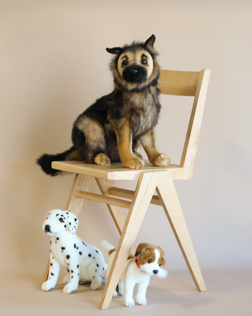 A realistic German Shepherd Puppy Stuffed Animal sits on a wooden chair, looking forward, with a toy Dalmatian and a small toy dog with a red collar standing next to it on the floor.