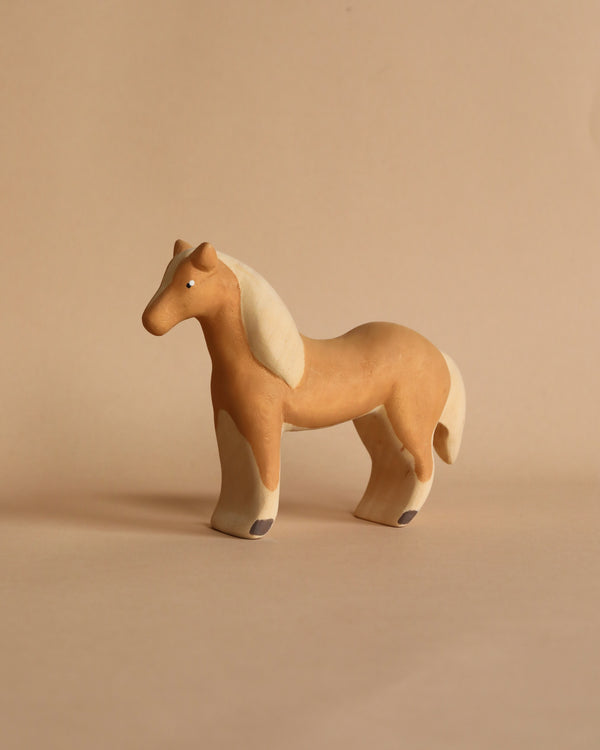 A small, light brown Handmade Wooden Horse with a white mane and tail stands on a neutral beige background. Handcrafted from linden wood, the horse has a simplistic and smooth design, giving it a minimalist and artistic appearance. Painted with child-safe paint, it's perfect for little ones.