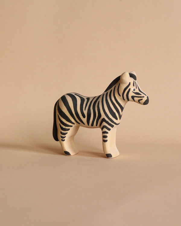 A small, Handmade Wooden Zebra with black stripes painted on its white body using non-toxic paint, standing on a beige surface. Handmade in Serbia, the background is also beige, creating a minimalist and unified color scheme.