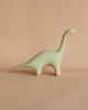 A small, handmade wooden Brachiosaurus dinosaur figurine with a long neck and tail, standing against a plain beige background.