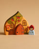 The Handmade Wooden Cottage set showcases a house with a rounded roof, beautifully adorned with a tree, windows, and an owl. Accompanying the house is a small figure dressed in white and red, reminiscent of Little Red Riding Hood. Perfect for imaginative play or as delightful decor, the backdrop features a solid beige color.