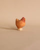 A small, handcrafted wooden figure of a chicken, made from linden wood with a simple, rounded shape, stands on a neutral beige background. The chicken is painted in natural tones with child-safe paint, offering a subtle contrast between the body and head.