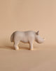 A small, light brown, wooden figurine of a rhinoceros stands on a beige background. The Handmade Wooden Rhinoceros from Meshka and Friends is carved simply with minimal details and features a slight smile, adding a whimsical feel to the artwork. Non-toxic paint ensures it's safe for all ages.