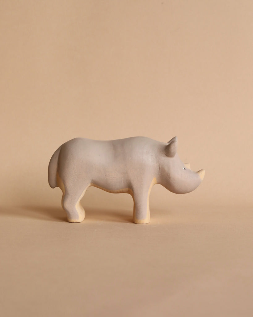 A small, light brown, wooden figurine of a rhinoceros stands on a beige background. The Handmade Wooden Rhinoceros from Meshka and Friends is carved simply with minimal details and features a slight smile, adding a whimsical feel to the artwork. Non-toxic paint ensures it's safe for all ages.